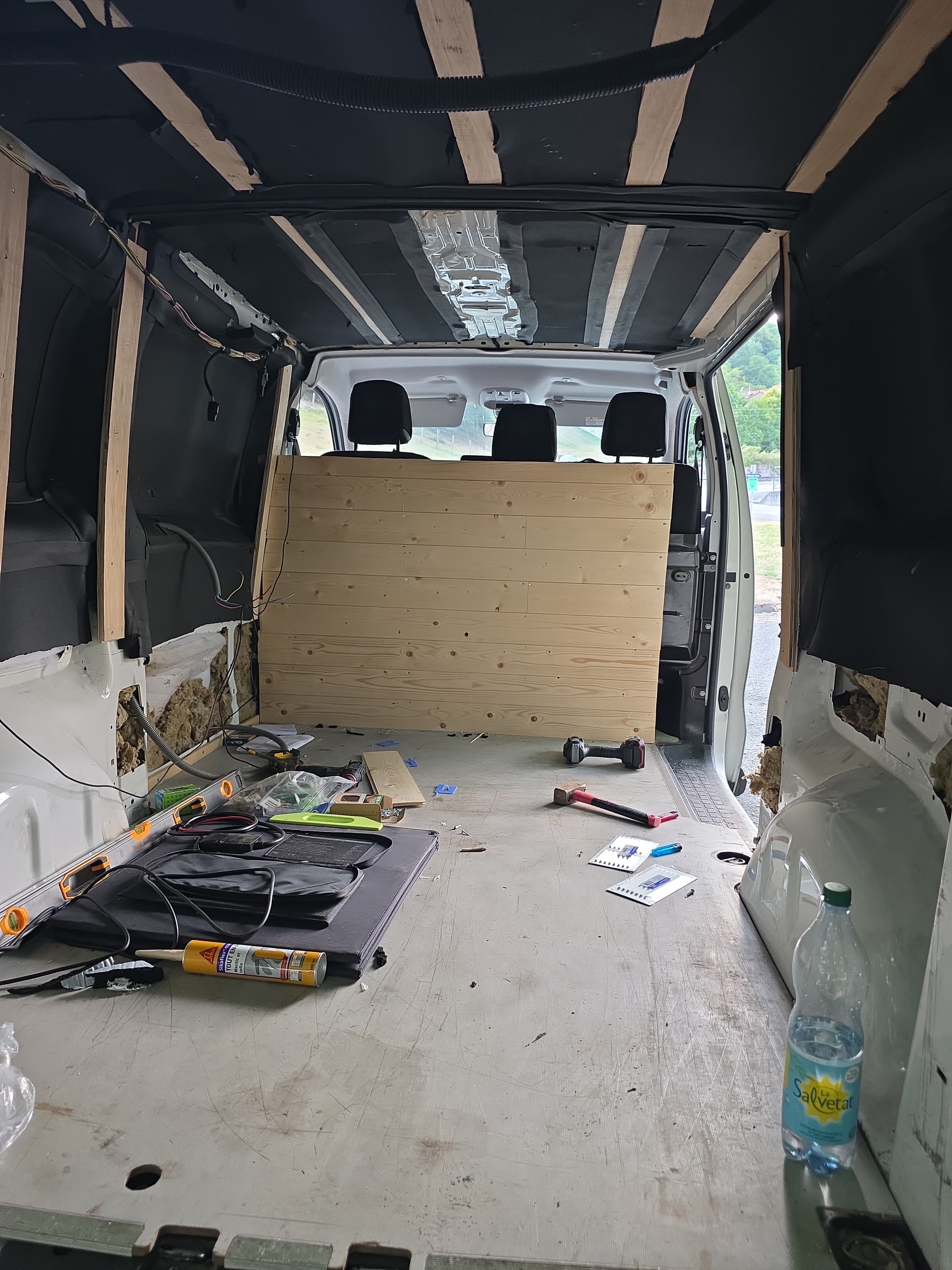 Aménagement intérieur d'un van en cours de réalisation, avec pose d'un lambris bois en cloison de séparation cabine/espace de vie, isolation en laine naturelle visible sur les parois latérales et début de pose de liteaux en plafond. Un travail de transformation artisanale soigné qui témoigne d'un vrai savoir-faire en menuiserie et aménagement intérieur sur-mesure.